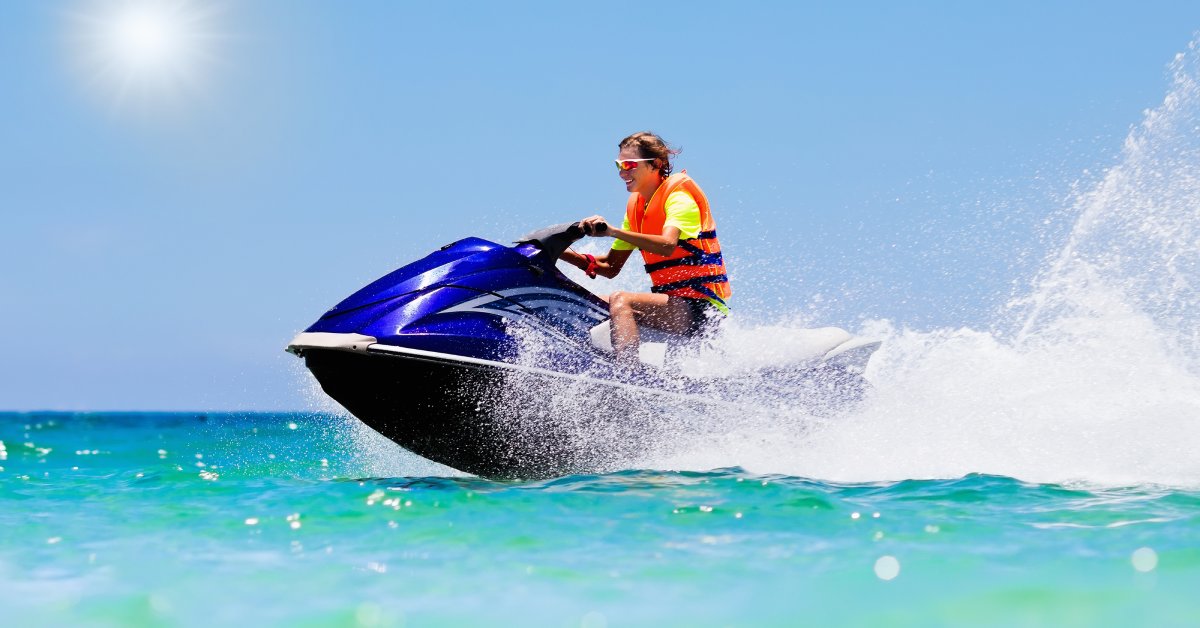 Ways To Extend the Life of Your Jet Ski Engine A young man in an orange life jacket and reflective sunglasses smiling while riding a blue jet ski on a sunny day.