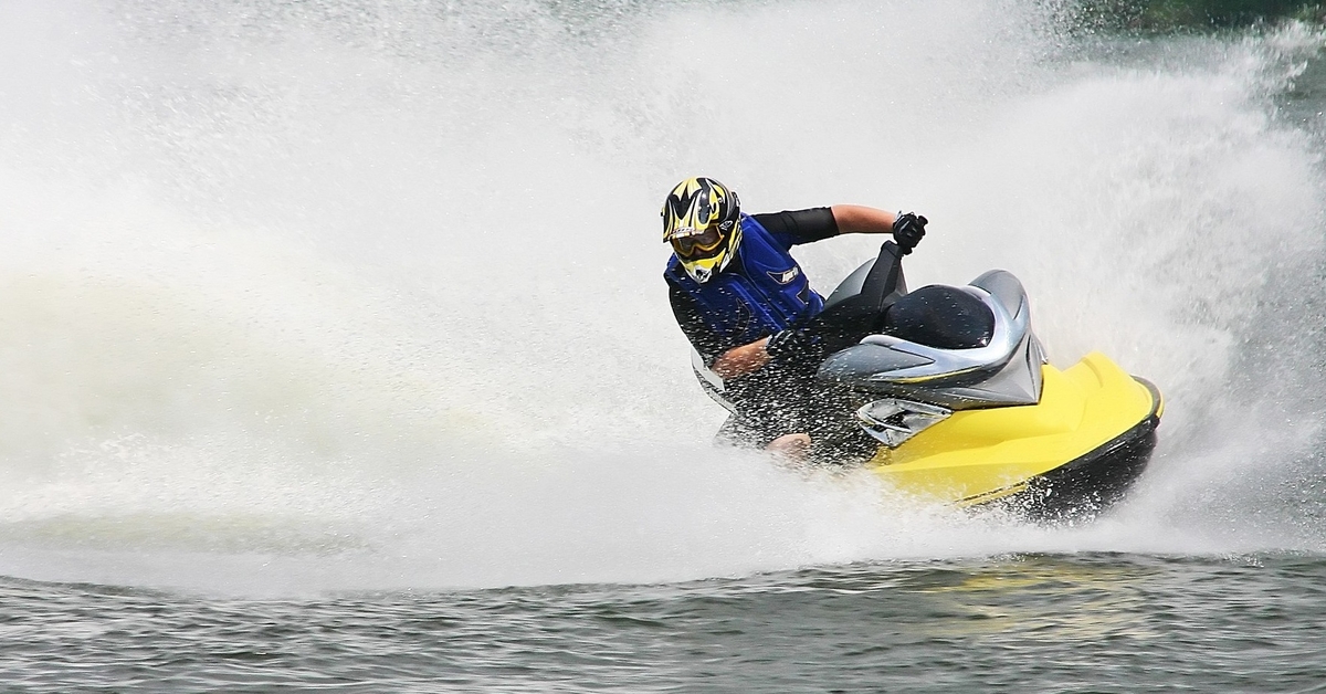 A person wearing a yellow helmet rides on a yellow jet ski on a large body of water, creating big waves in their wake.