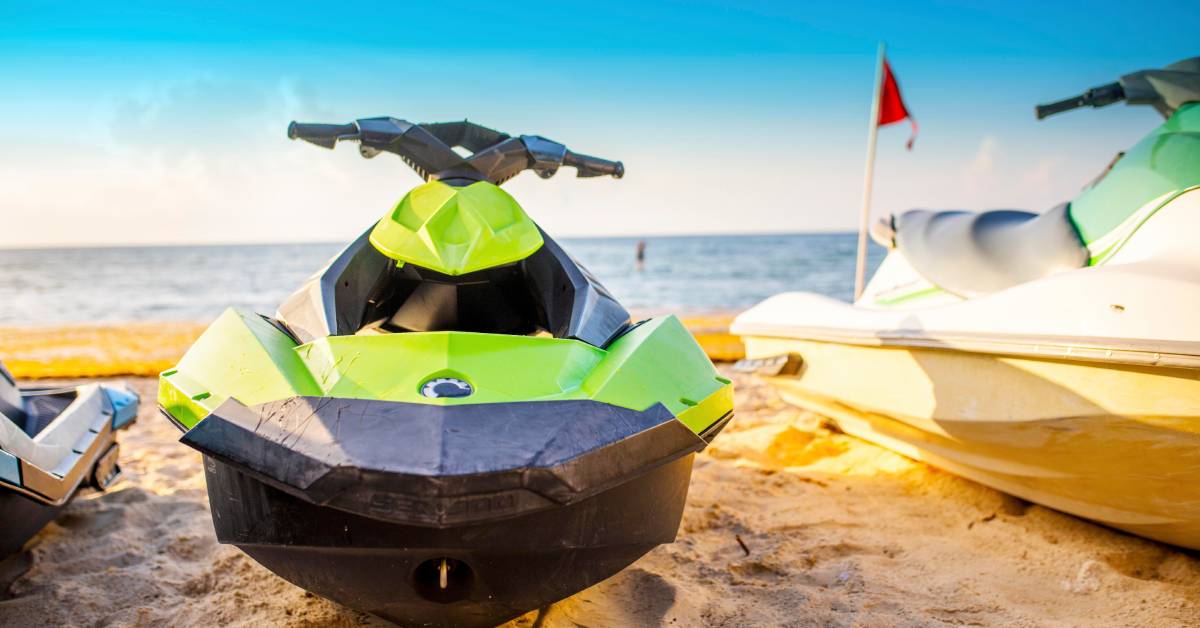 A green jet ski is on the sand with the water behind it. The sun shines down and there are water drips on the jet ski.
