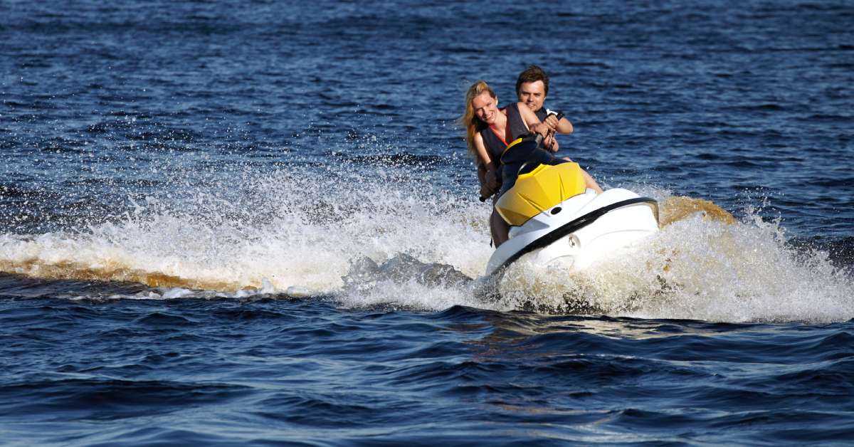 A couple wearing lifejackets rides a yellow and white jet ski across the deep blue water with their wake in the background.