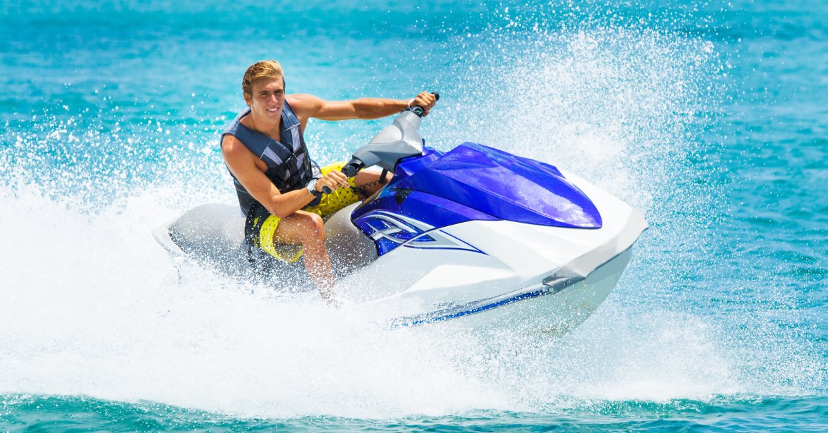 A young man rides on a big jet ski, cruising over and creating blue waves, leaving mist in their wake.