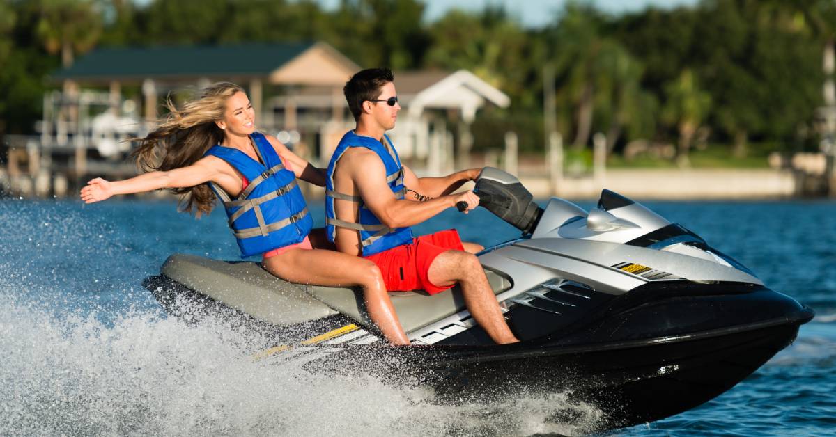 A man and a woman ride on the back of a jet ski, splashing water. In the background there are trees and some homes.