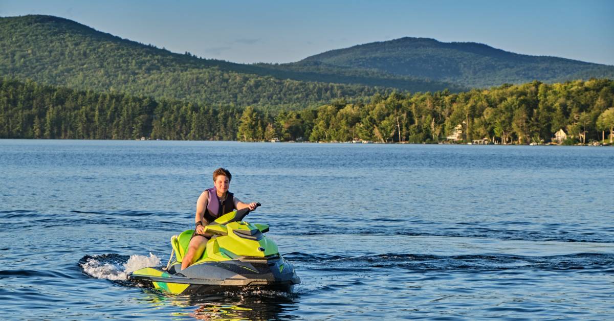 A person rides a green jet ski on a still lake with a huge forest of trees and hills behind them in the distance.