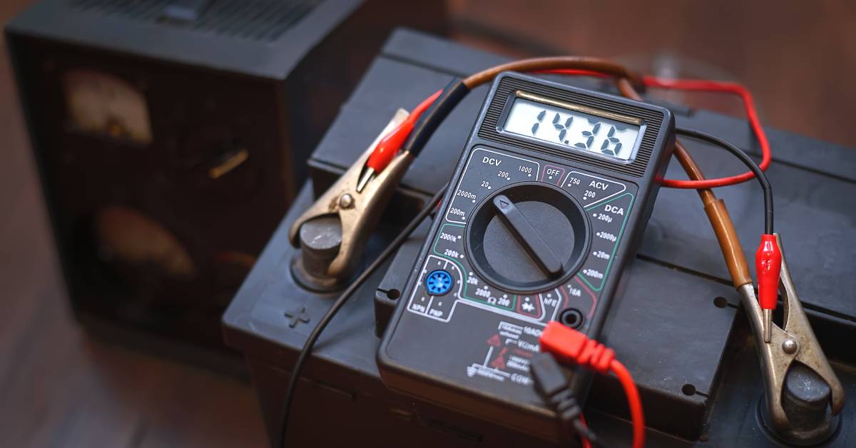 A multimeter is checking the charge of a battery with clamps on positive and negative nodes, reading 14.36 on a screen.